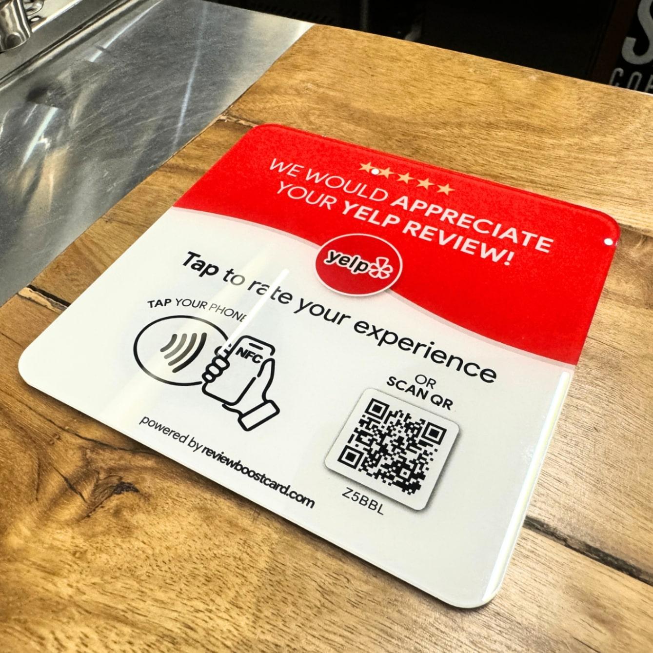 A ReviewBoost plaque for Yelp reviews on a wooden counter, featuring an NFC tap area and QR code for easy review submission
