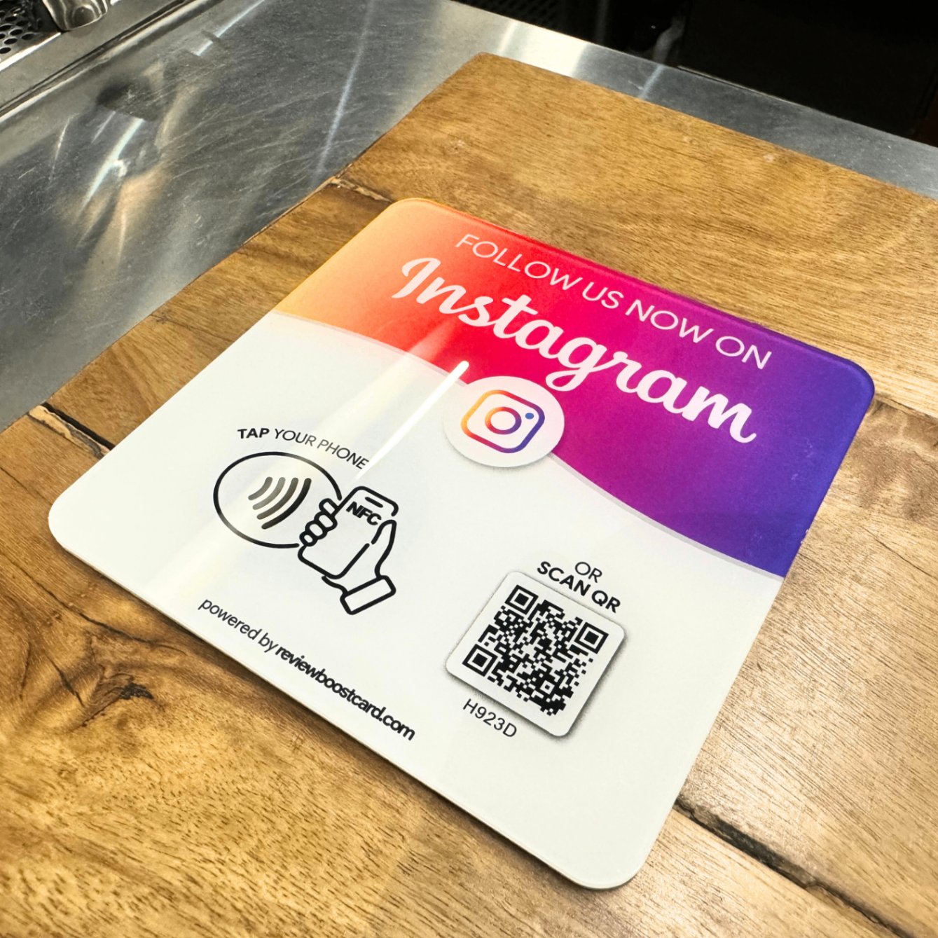 A square plaque with a gradient from orange to purple, featuring the Instagram logo and text "Follow Us Now on Instagram." It offers options to "Tap Your Phone" using NFC or "Scan QR" code, powered by reviewboostcard.com, placed on a wooden surface