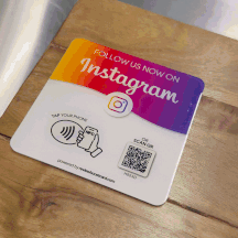 An animated GIF showing an Instagram follow plaque on a wooden surface. The plaque has a gradient from orange to purple, featuring the Instagram logo, with instructions to "Tap Your Phone" using NFC or "Scan QR" code