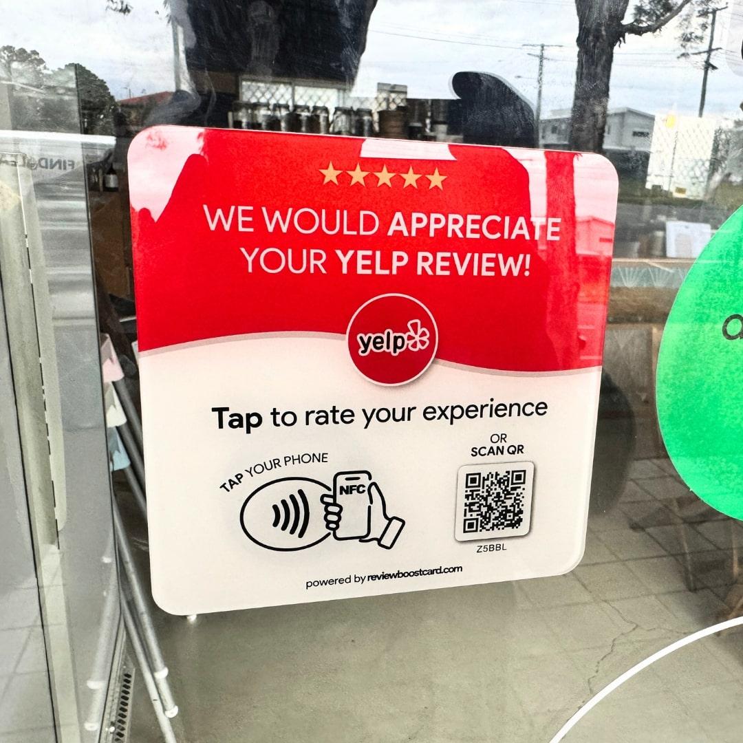 A ReviewBoost plaque requesting Yelp reviews displayed on a glass door, featuring an NFC tap area and QR code for easy review submission